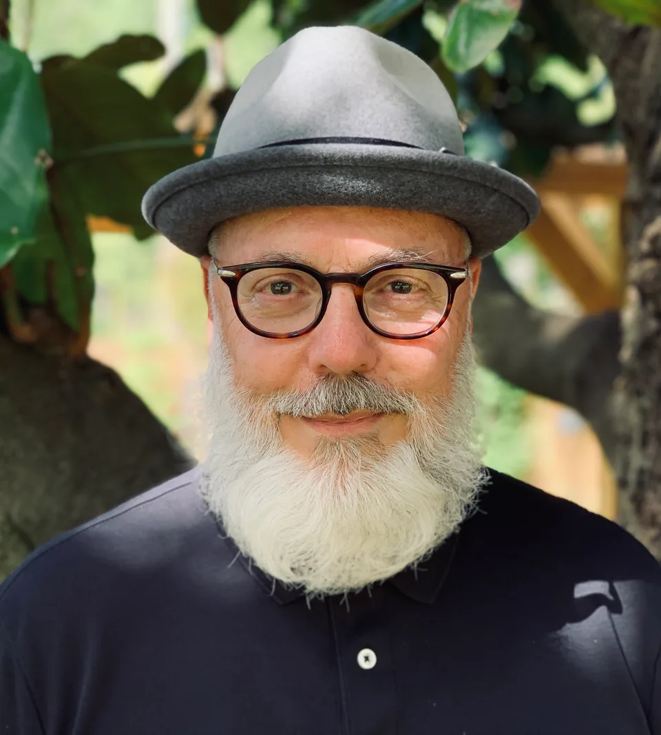 Rabbi Rami, smiling, with a white beard, round glasses, and a grey hat, among green trees